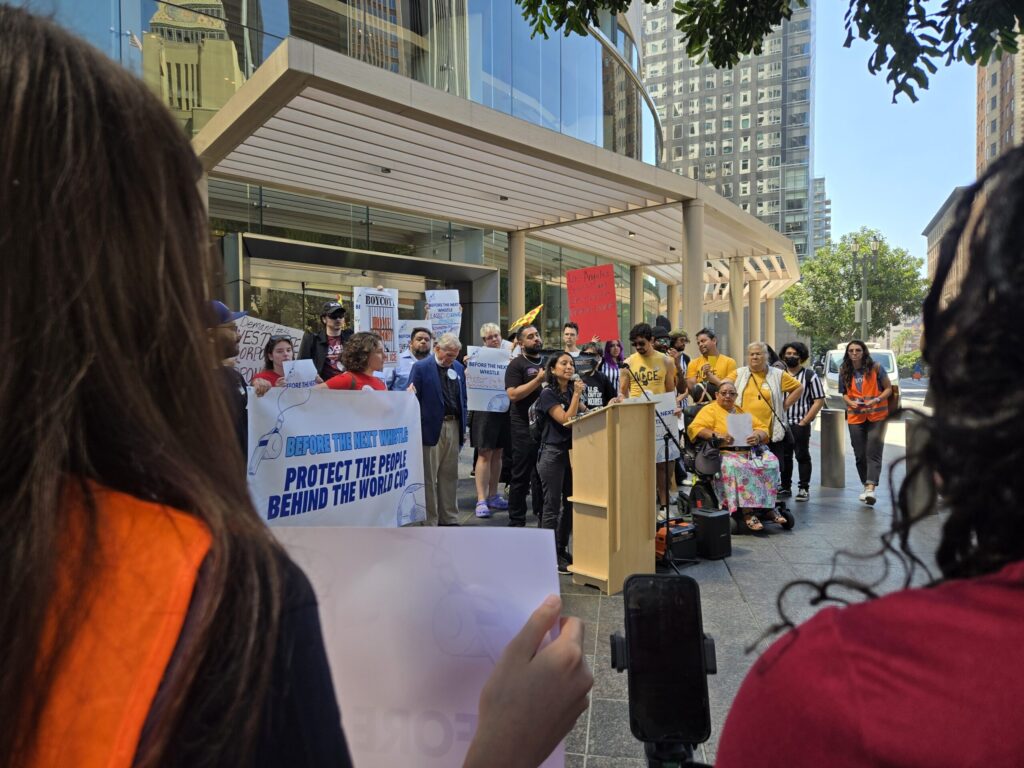20260709_103030 - Jobs to Move America Crowd of diverse protesters holding signs outside a glass-fronted building, with a speaker at a podium in the foreground.