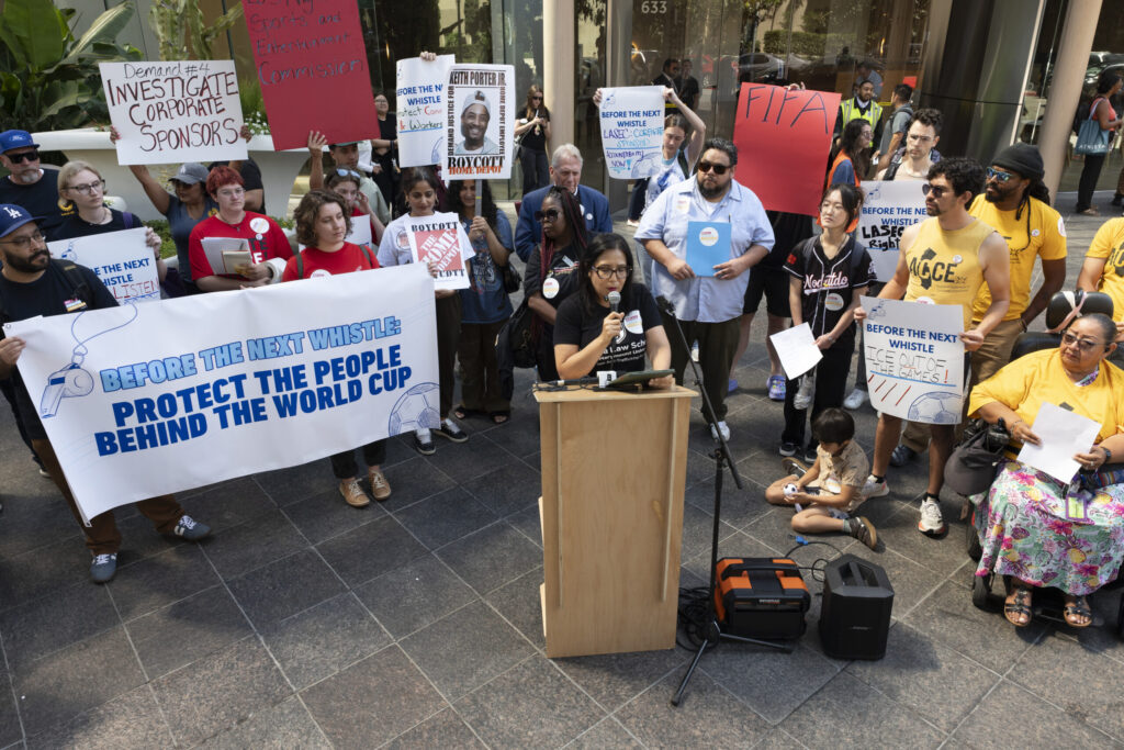 Diverse group of protesters outside a building, holding signs and banners urging accountability and whistleblower protection, with a speaker at a wooden podium addressing the crowd.